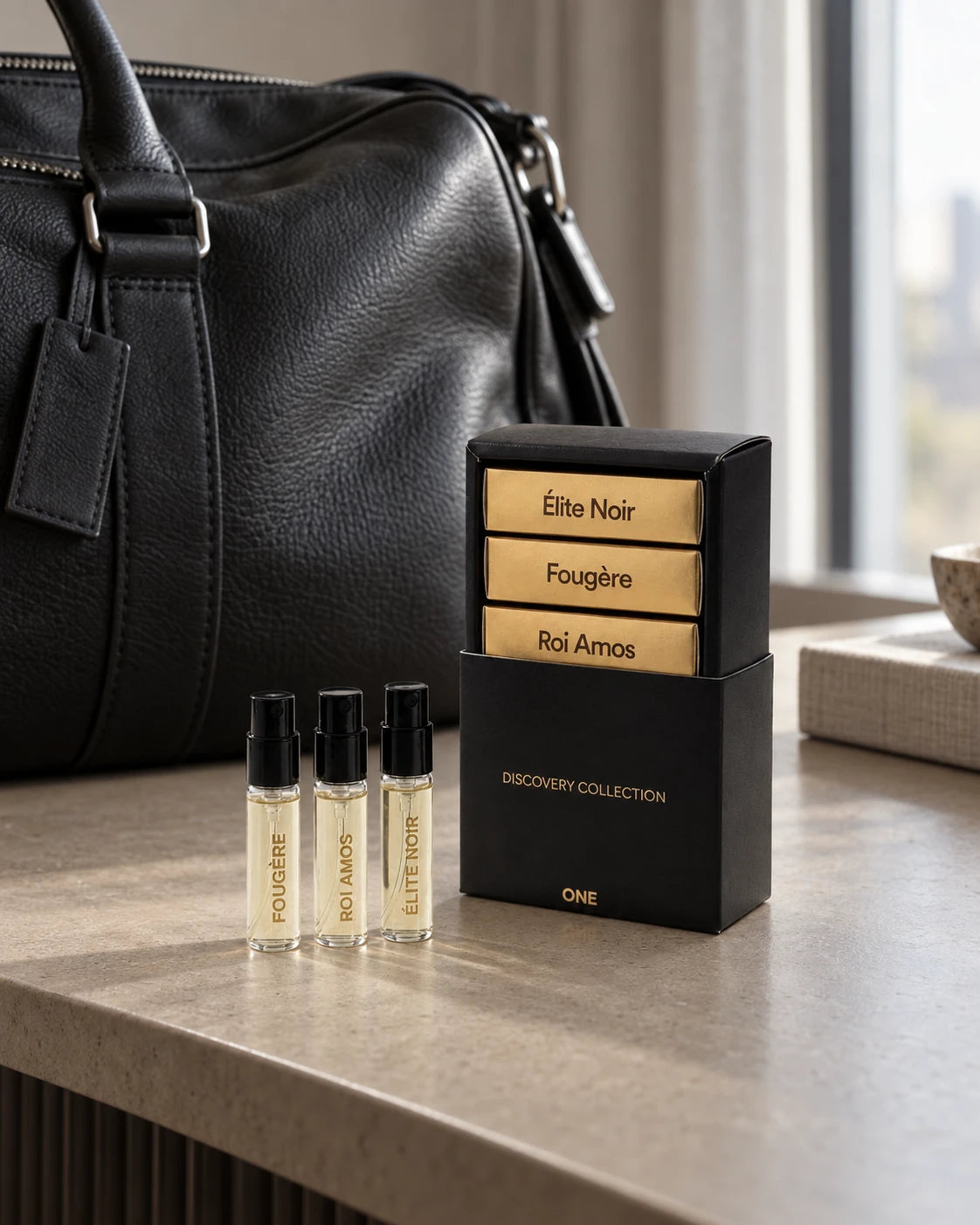 ONE Discovery Collection with three 3 ml signature fragrance sprays and its black-and-gold presentation sleeve beside a leather weekender bag